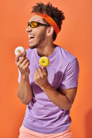 young african american man in sunglasses holding bite-sized donuts and laughing on orange background