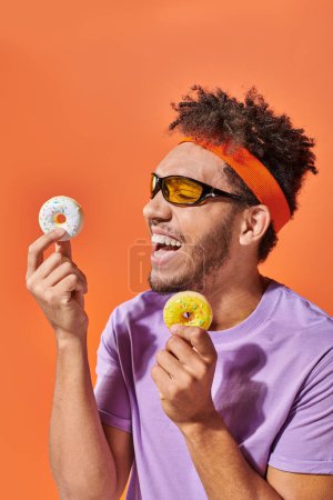fröhlicher afrikanisch-amerikanischer Mann mit Sonnenbrille, mundgerechte Donuts in der Hand und lachend auf orangefarbenem Hintergrund