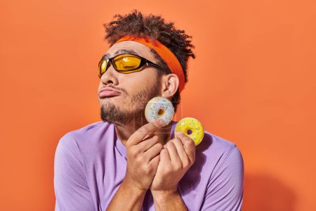 Foto de Hombre afroamericano en gafas de sol sosteniendo donuts tamaño mordisco sobre fondo naranja, mueca - Imagen libre de derechos
