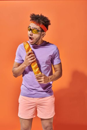 funny african american man in sunglasses and headband biting fresh baguette on orange background