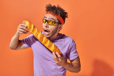 Afrikanisch-amerikanischer Mann mit Sonnenbrille und Stirnband beißt frisches Baguette auf orangefarbenem Hintergrund, Bäckerei
