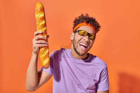 happy african american man in sunglasses and headband holding fresh baguette on orange background