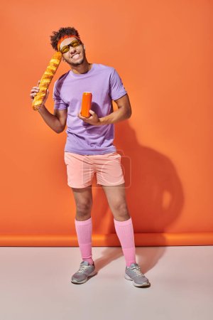 cheerful african american man in sunglasses holding fresh baguette and soda on orange background