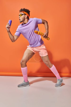 young african american man in sunglasses holding fresh baguette and soda on orange background