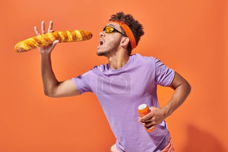 hombre afroamericano apasionado en gafas de sol comiendo baguette fresca y sosteniendo soda en naranja