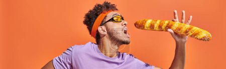 banner, african american man in sunglasses and headband holding fresh baguette on orange background