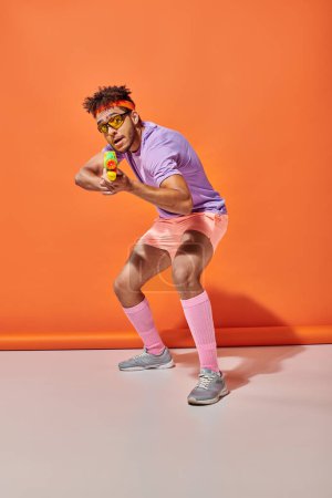 young african american guy in sunglasses holding water gun on orange background, summer fun