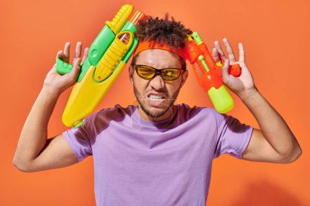 hombre afroamericano tenso en gafas de sol con pistolas de agua sobre fondo naranja, juguetón