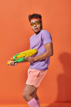 Foto de Hombre afroamericano confuso en gafas de sol sosteniendo pistola de agua sobre fondo naranja, mueca - Imagen libre de derechos