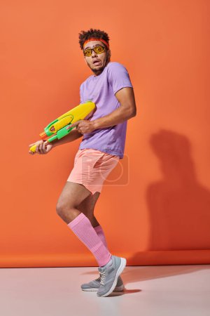 surprised african american man in sunglasses holding water gun on orange background, grimace