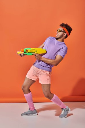 tendu afro-américain mec dans des lunettes de soleil jouer combat de l'eau avec pistolet jouet sur fond orange