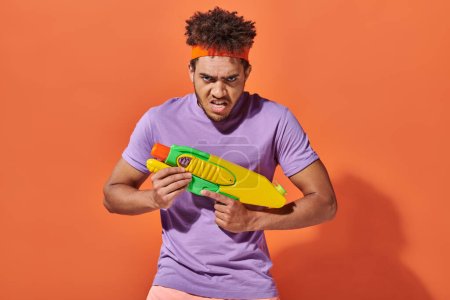 african american man in headband playing water fight with toy gun on orange background, grimace
