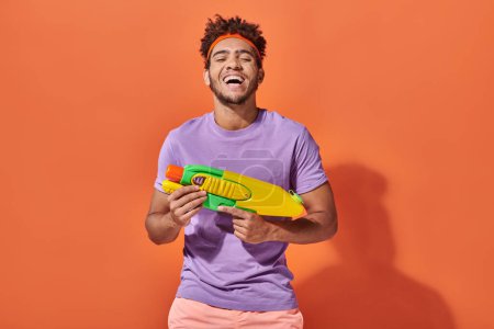 glücklich afrikanisch-amerikanischer Mann im Stirnband spielt Wasserschlacht mit Spielzeugpistole auf orangefarbenem Hintergrund