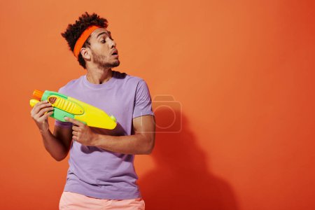 curly african american man in headband playing water fight with toy gun on orange background