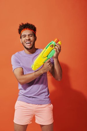positiver afrikanisch-amerikanischer Mann im Stirnband spielt Wasserschlacht mit Spielzeugpistole auf orangefarbenem Hintergrund