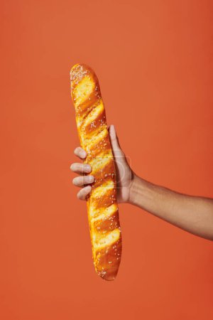 vista recortada de la persona que sostiene la baguette recién horneada sobre fondo naranja, panadería francesa crujiente