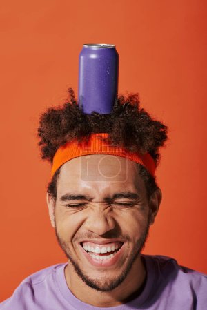 lila Getränkedose auf dem Kopf des glücklichen lockigen afrikanisch-amerikanischen Typen mit Stirnband auf orangefarbenem Hintergrund