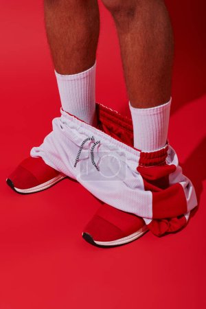 Foto de Foto conceptual, hombre recortado en zapatillas de deporte, calcetines blancos y corredores de pie sobre fondo rojo - Imagen libre de derechos