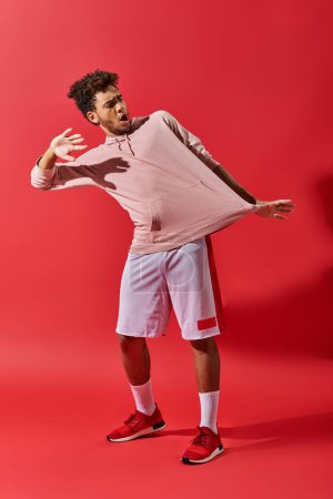 Foto de Sorprendido hombre afroamericano mirando ropa de gimnasio y de pie con la boca abierta sobre fondo rojo - Imagen libre de derechos