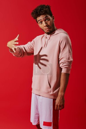 confused african american man in hoodie gesturing and looking at camera on red background, shadow