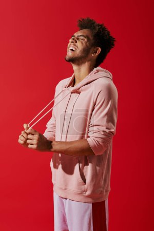 happy african american man pulling drawstrings of pink hoodie and laughing on red background