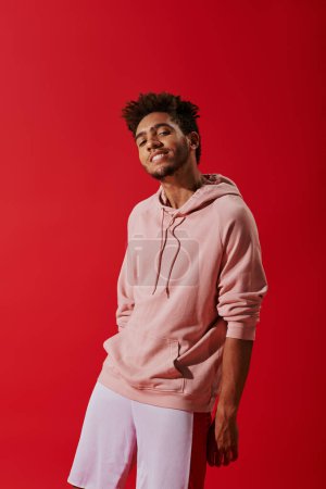 happy african american man in pink hoodie looking at camera while smiling on red background