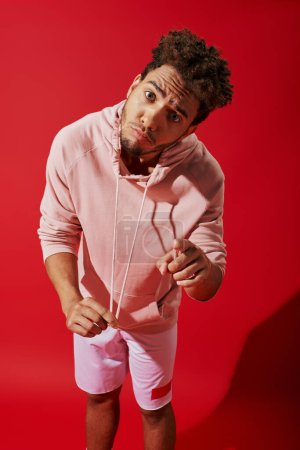 african american man pulling drawstrings of pink hoodie and looking at camera on red background Poster 692588020