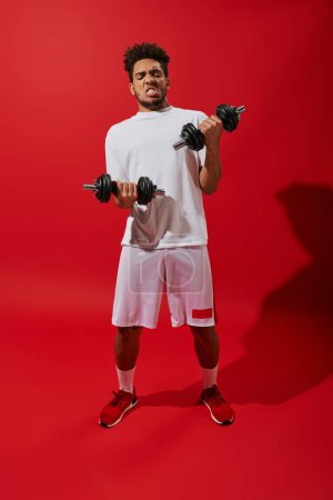 curly african american sportsman working out with heavy dumbbells on red background, grimace