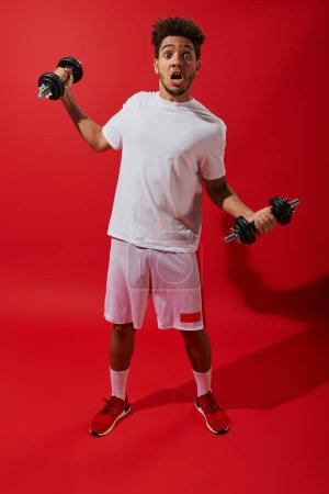 shocked african american sportsman working out with heavy dumbbells on red background, grimace
