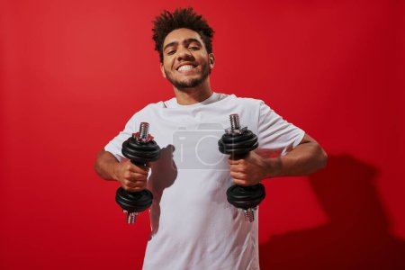 fröhlicher afrikanisch-amerikanischer Sportler beim Training mit schweren Hanteln auf rotem Hintergrund, Power