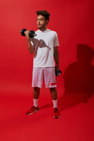 longitud completa de deportista afroamericano haciendo ejercicio con pesadas mancuernas sobre fondo rojo, poder
