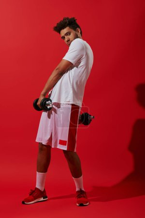 longitud completa de deportista afroamericano ejerciendo con pesadas mancuernas sobre fondo rojo, poder