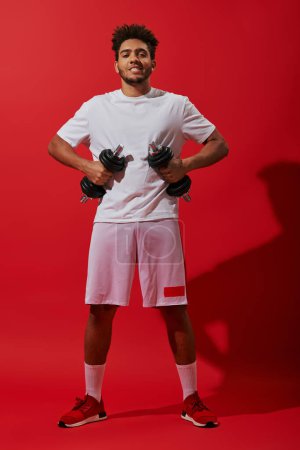 full length of african american sportsman exercising with dumbbells on red background, powerful