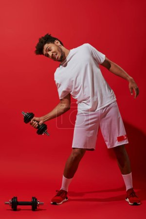 longitud completa del hombre americano africano divertido en ropa deportiva que trabaja hacia fuera con la mancuerna sobre fondo rojo