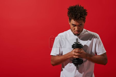 african american man in sportswear working out with dumbbell and looking at camera on red background