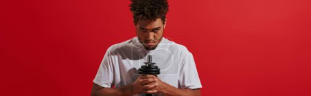 african american man in sportswear working out with dumbbell and looking at camera on red, banner