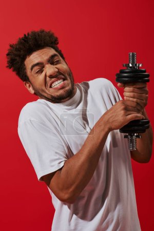 joven y rizado afroamericano deportista haciendo ejercicio con pesada mancuerna sobre fondo rojo