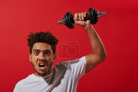 motivierter afrikanisch-amerikanischer Mann in Sportbekleidung beim Training mit schwerer Hantel auf rotem Hintergrund
