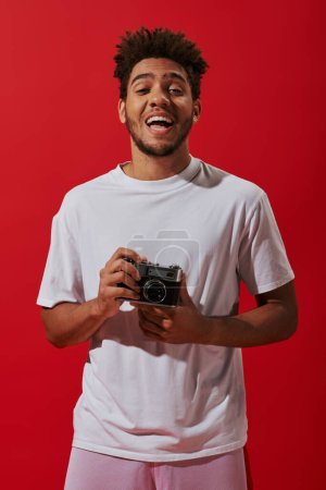 happy african american photographer holding vintage camera and smiling on red background, hobby