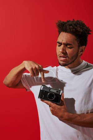 young african american man looking at his vintage camera on red background, pushing button