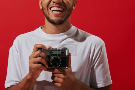 Foto de Recortado y feliz afroamericano fotógrafo celebración vintage cámara y sonriendo sobre fondo rojo - Imagen libre de derechos