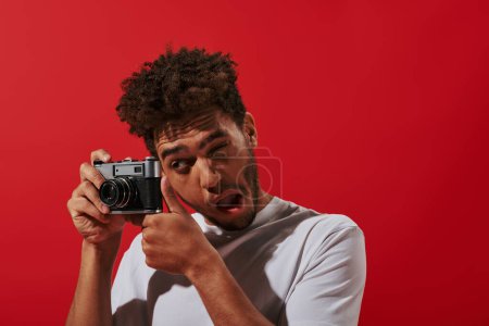 divertido fotógrafo afroamericano mirando al visor mientras toma fotos sobre fondo rojo