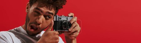 Téléchargez les photos : Drôle homme afro-américain regardant viseur tout en prenant des photos sur fond rouge, bannière - en image libre de droit
