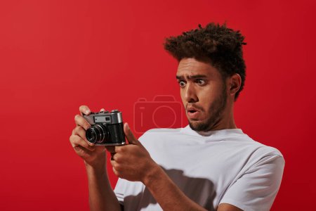 Photo for Funny african american photographer looking at retro camera while taking shot on camera on red - Royalty Free Image