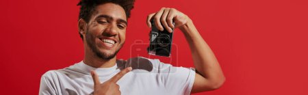 Photo for Happy african american photographer pointing at retro camera and smiling on red background, banner - Royalty Free Image