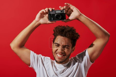 joyeux photographe afro-américain tenant un appareil photo rétro et souriant sur fond rouge