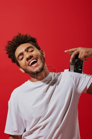 optimistic african american photographer taking shot on retro camera and smiling on red background