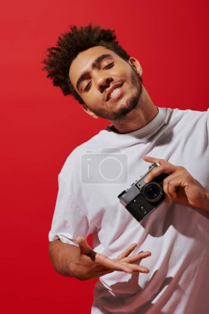 young african american man taking shot on retro camera and smiling on red background, creative