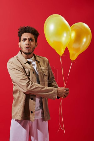 shocked african american man in beige jacket holding helium balloons on red background, party