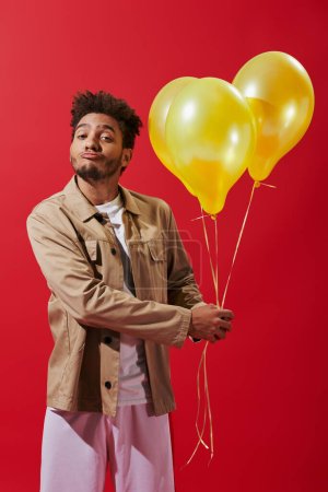 funny african american man in beige jacket holding helium balloons on red background, party